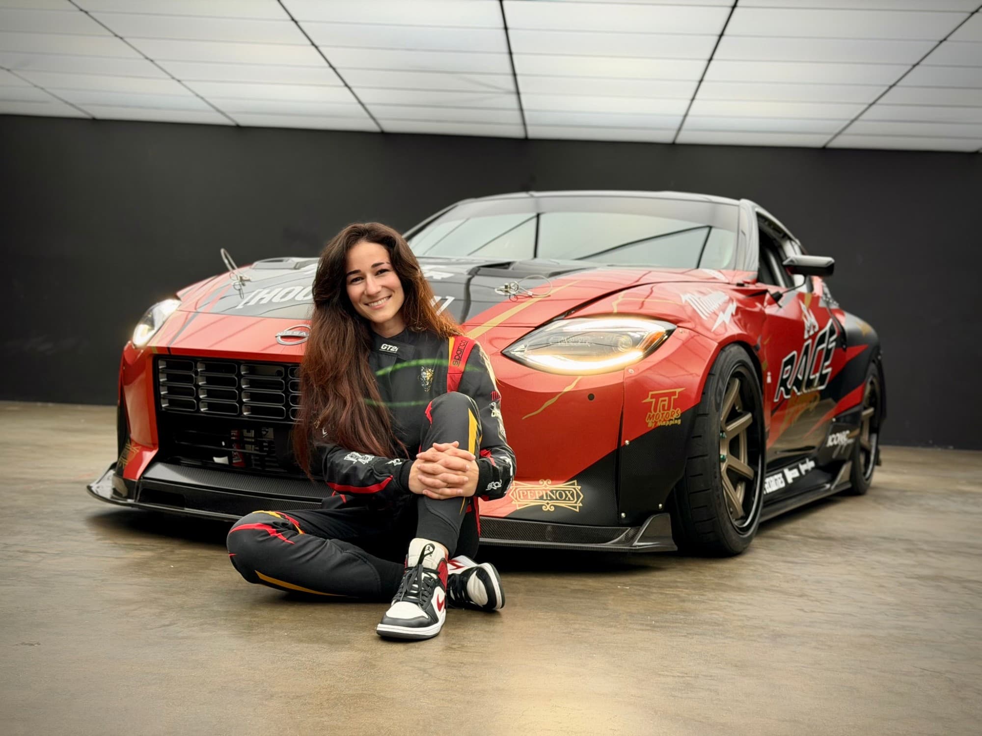 Pilote de drift assise devant la Nissan 400Z à la livrée rouge, noire et or couverte de sponsors, dans un studio